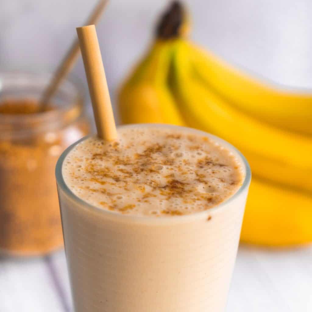 A close-up of a thick and creamy vanilla banana protein smoothie in a glass with a bamboo straw, dusted with cinnamon next to fresh yellow bananas.
