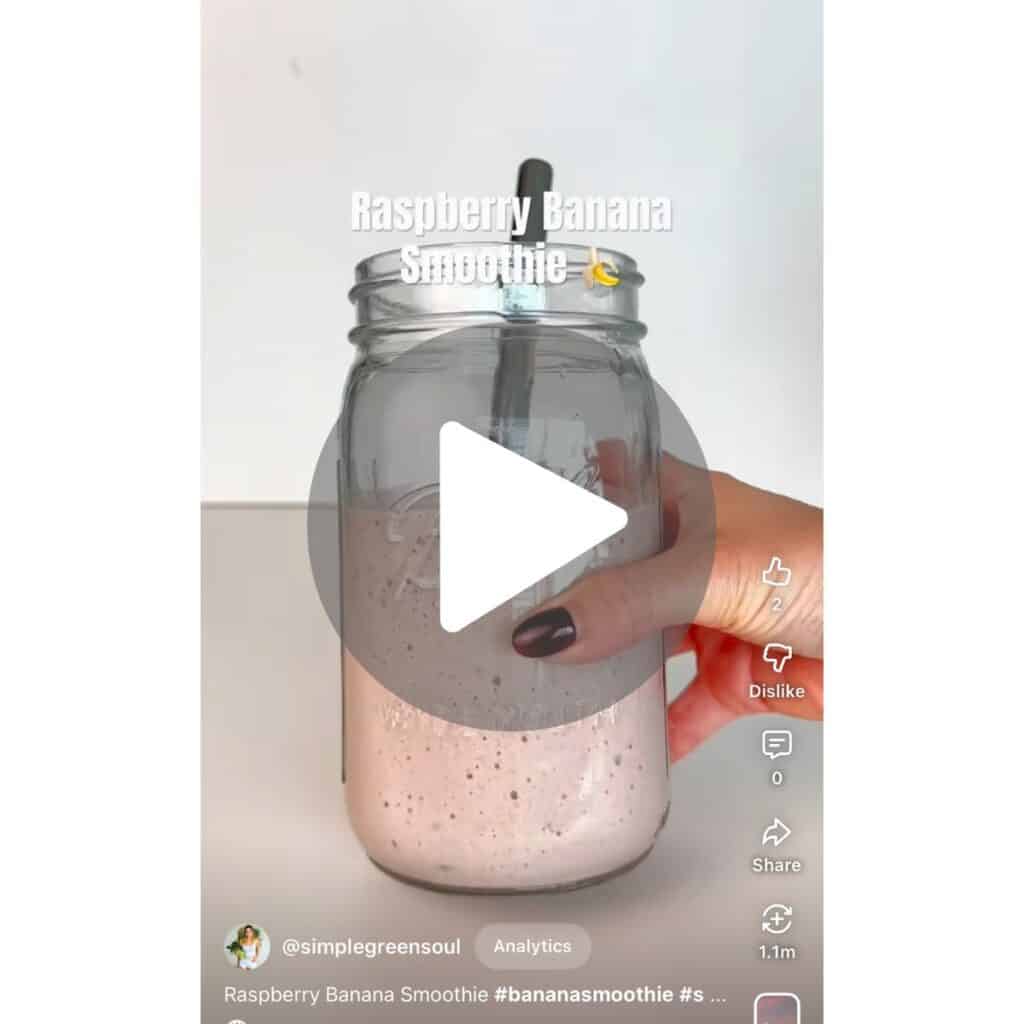 A YouTube Shorts video screenshot of a vibrant pink raspberry banana smoothie being made in a blender, with a link to watch the full recipe tutorial.
