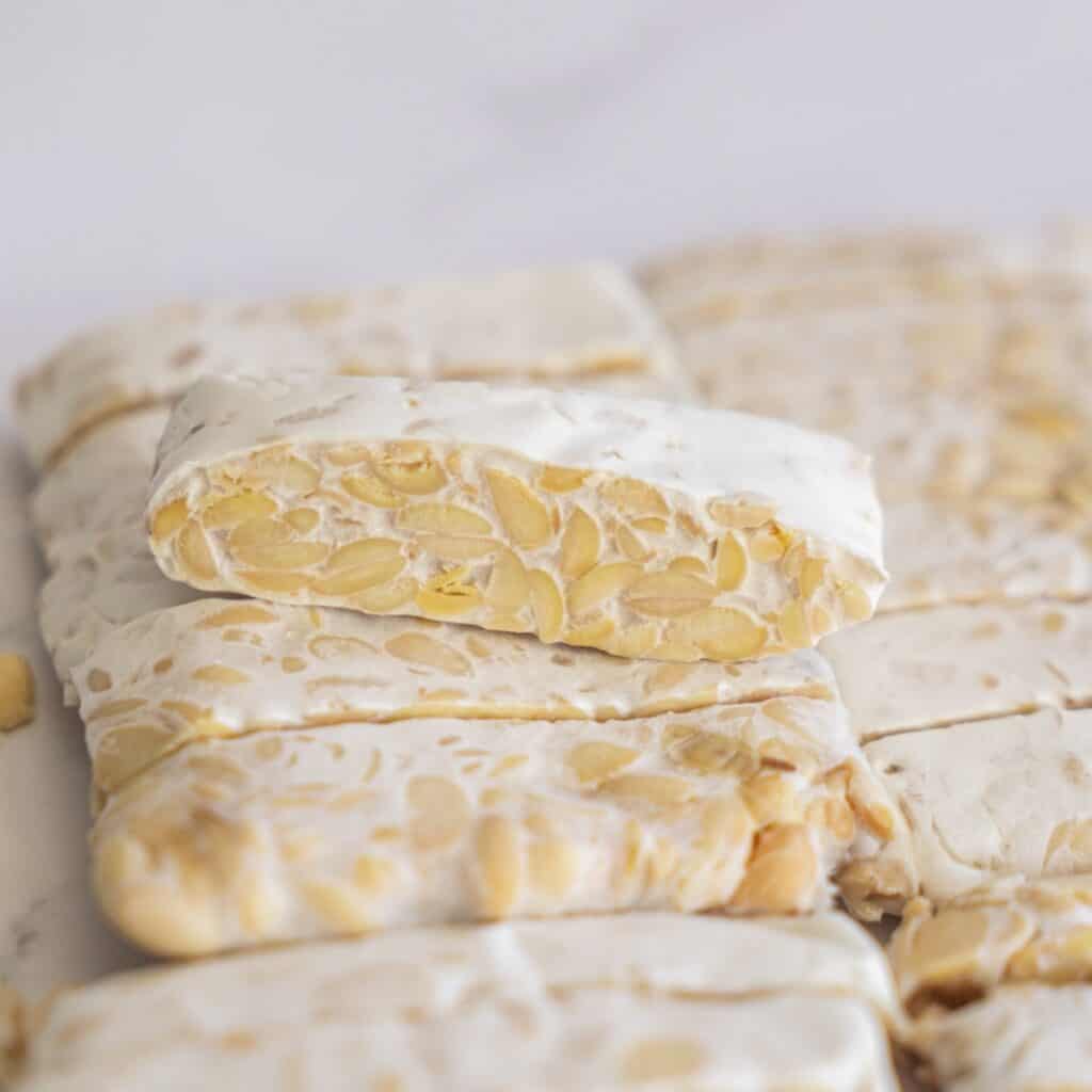 Close-up of raw, uncooked blocks of tempeh stacked on top of each other, showing the texture of the fermented whole soybeans.