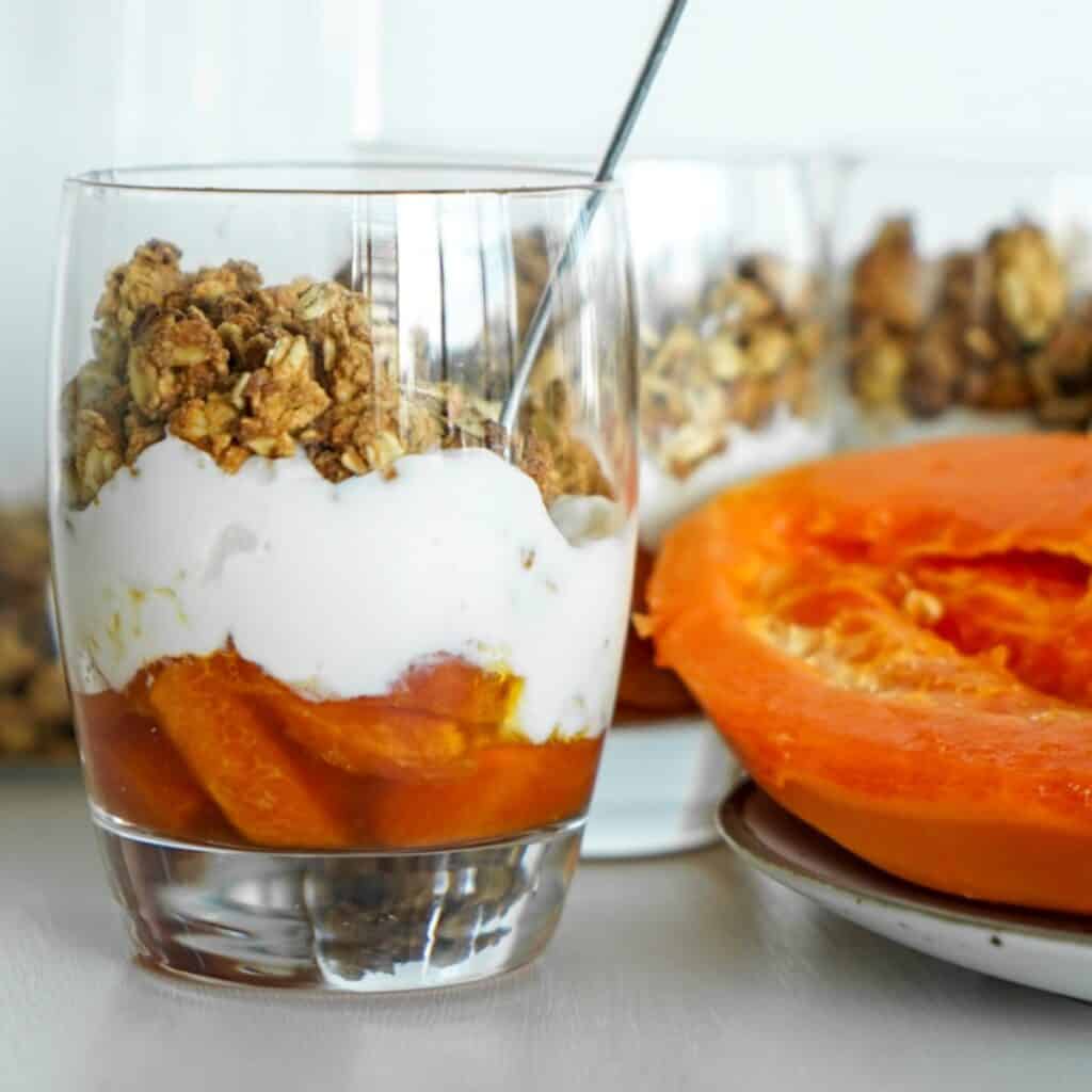 A vibrant granola cups recipe featuring warm baked papaya, plant-based coconut yogurt, and crunchy high-protein oatmeal granola in a clear glass meal prep cup.