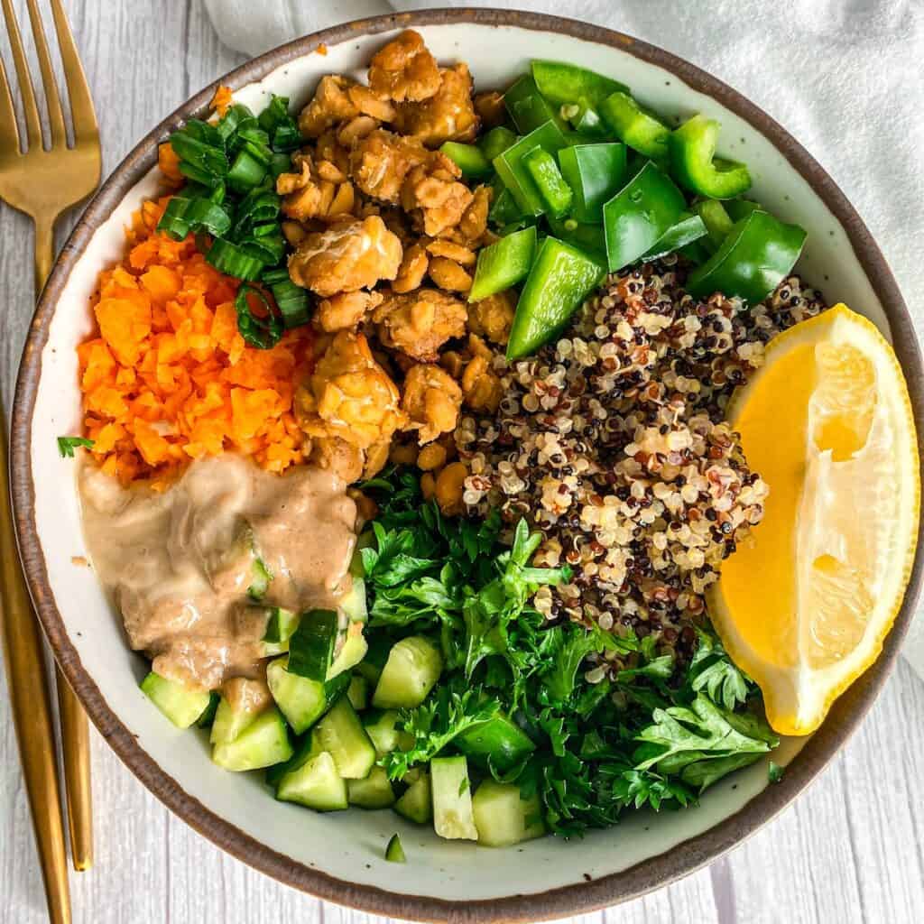 Vibrant high-protein tempeh salad bowl with quinoa, grated carrots, diced cucumbers, and green capsicum, drizzled with a creamy vegan tahini dressing.