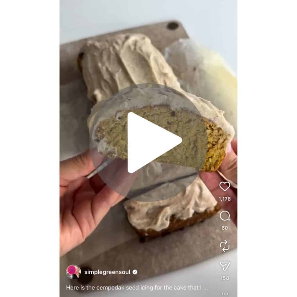 A screenshot of a viral Instagram video showing a close-up of a hand holding a freshly sliced piece of healthy cempedak cake topped with thick vanilla seed icing