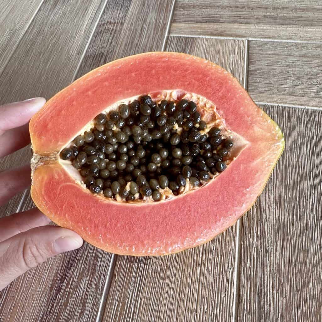 A hand holding a ripe papaya, sliced in half to show the orange flesh and black seeds in the centre