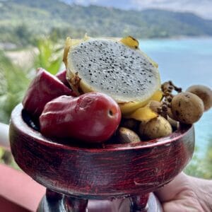 A dark wooden bowl filled with fresh exotic Asian fruits, including sliced red dragon fruit, rambutans, and mangosteens, with a tropical Malaysian beach in the background