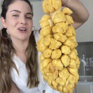 A close-up of a person holding a completely peeled cempedak fruit up by its central stem in the kitchen, showcasing the massive cluster of ripe golden fruit pods.
