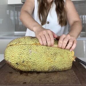 A close-up of a person using their fingernails to gently pry and peel the tough outer rind away from the inner cempedak fruit flesh.