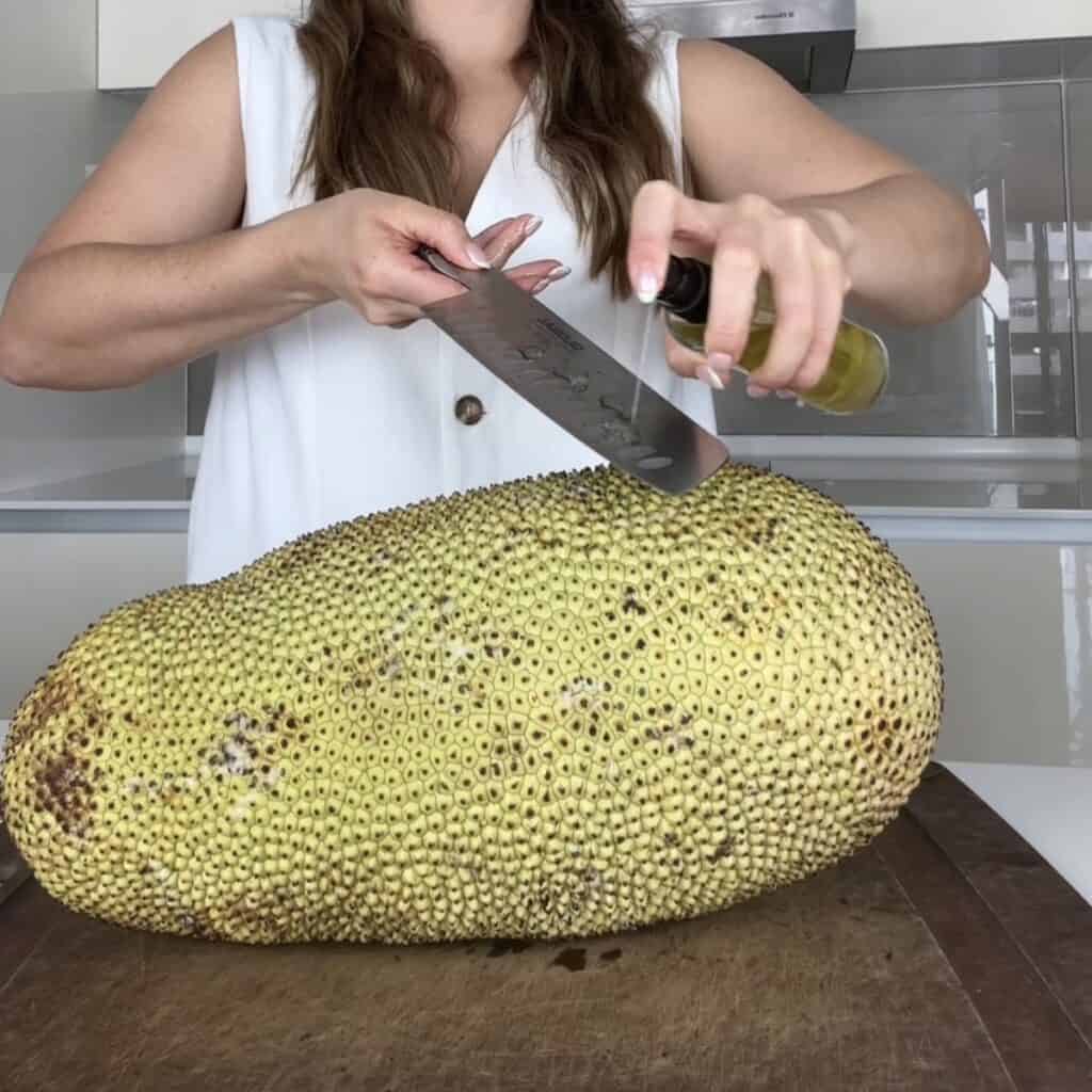 A close-up of a person coating a large kitchen knife with cooking oil over a whole cempedak fruit to prevent sticky latex sap from sticking to the blade.