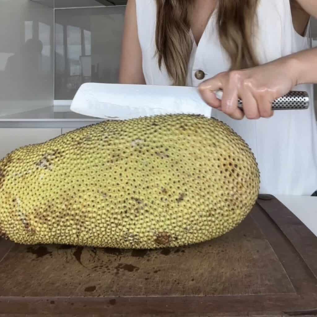 Cutting a cempedak fruit in half lengthwise with an oiled knife, scoring only the thick outer rind without slicing the flesh inside.