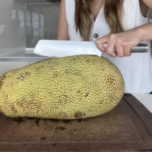 A close-up of a person coating a large kitchen knife with cooking oil over a whole cempedak fruit to prevent sticky latex sap from sticking to the blade.