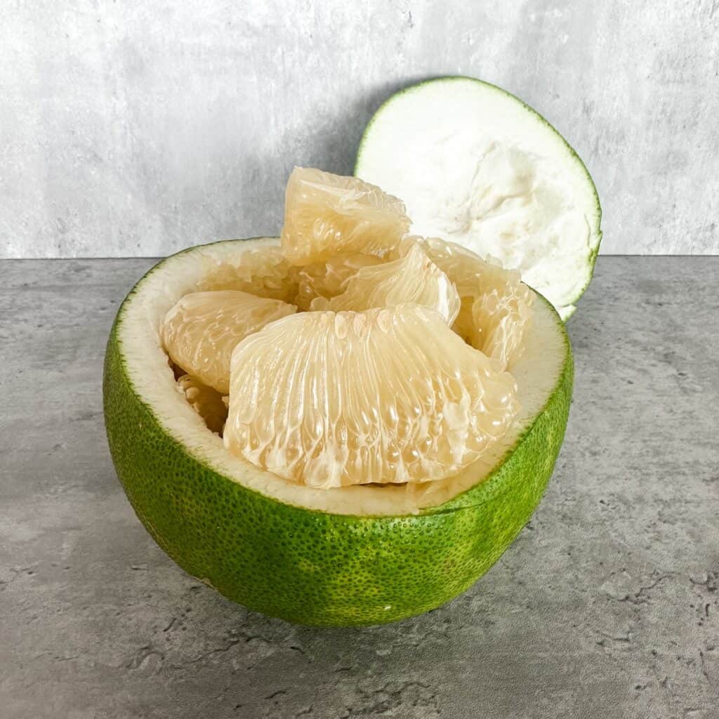 A green pomelo cut in half horizontally on a grey countertop. The bottom half serves as a natural bowl filled with peeled, juicy, light yellow pomelo segments, while the top half rests in the background showing its thick white pith.
