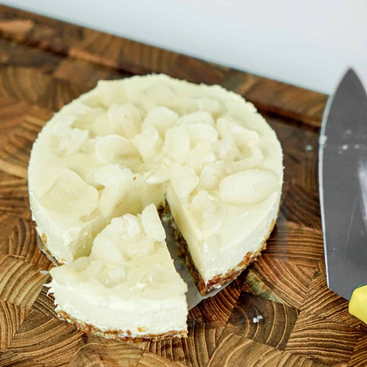 A top-down view of a sliced, no-bake vegan rambutan cheesecake on a wooden cutting board, showcasing its creamy tropical filling and crumbly date-cashew crust.