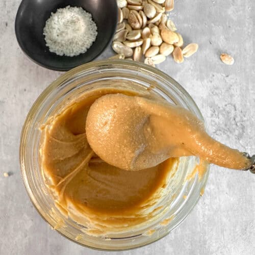 Smooth homemade Vitamix peanut butter in a glass jar with a spoon, roasted peanuts and a pinch of salt.