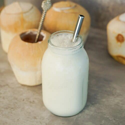 Creamy coconut shake made with fresh coconut water and coconut meat served in a mason jar with young coconuts in the background.